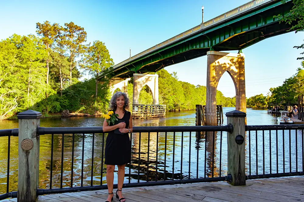 Woman with yellow flowers by river bridge.